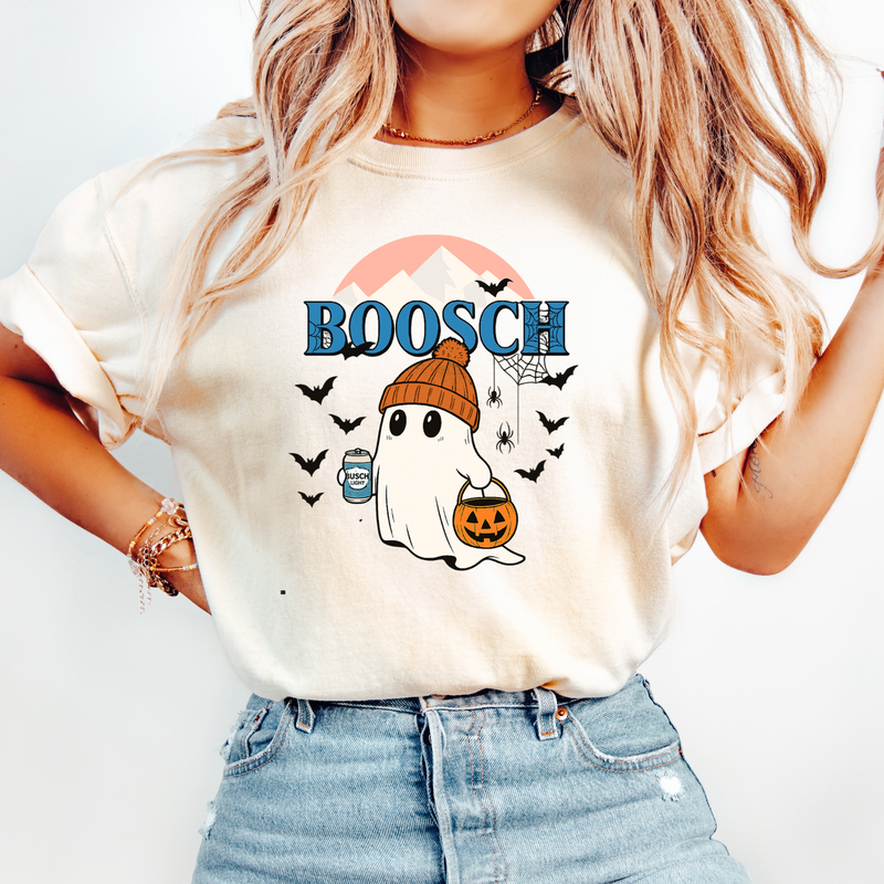 A woman wearing a cream-colored graphic t-shirt with a Halloween design that reads 'BOOSCH' in blue letters above a cartoon ghost in a brown beanie holding a jack-o'-lantern, paired with light blue denim jeans.
