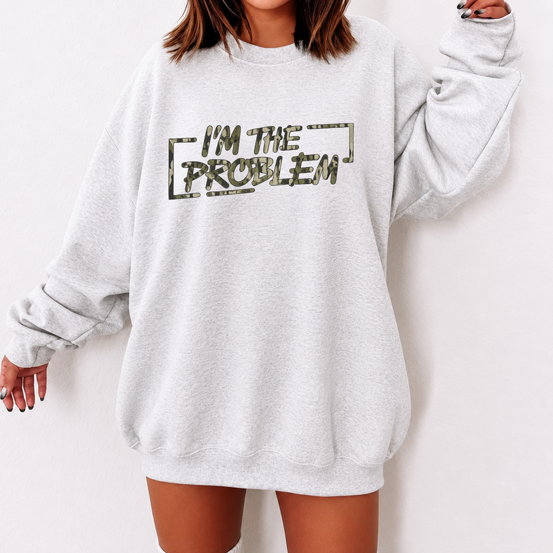 A woman wearing an oversized light gray sweatshirt with a distressed graphic that reads 'I'M THE PROBLEM' in dark olive lettering, shown against a plain light background.