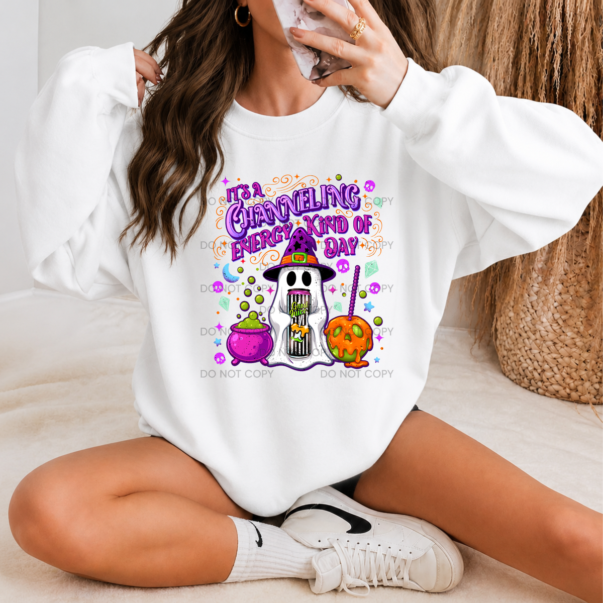 A woman sitting cross-legged wearing a white sweatshirt with a colorful Halloween graphic that reads 'IT'S A CRAZY HELLING EVERY KIND OF DAY' above cartoon ghosts and pumpkins. She is also wearing white socks and white sneakers, with a neutral indoor background and a woven basket.