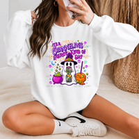 A woman sitting cross-legged wearing a white sweatshirt with a colorful Halloween graphic that reads 'IT'S A CRAZY HELLING EVERY KIND OF DAY' above cartoon ghosts and pumpkins. She is also wearing white socks and white sneakers, with a neutral indoor background and a woven basket.