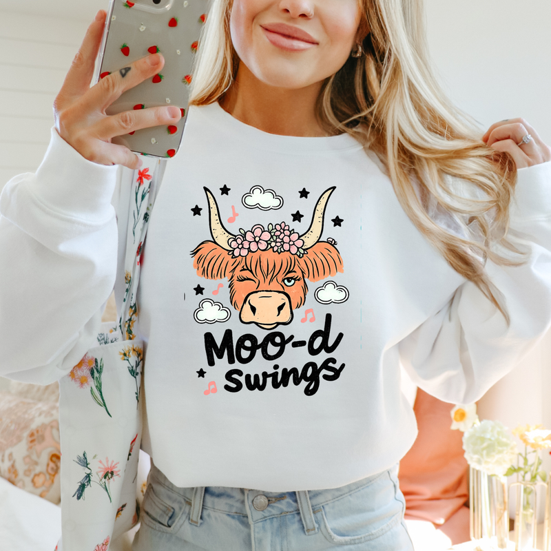 A woman takes a mirror selfie wearing a white crewneck sweatshirt with a graphic of a horned cow wearing a flower crown and the text 'Moo-d Swings'.