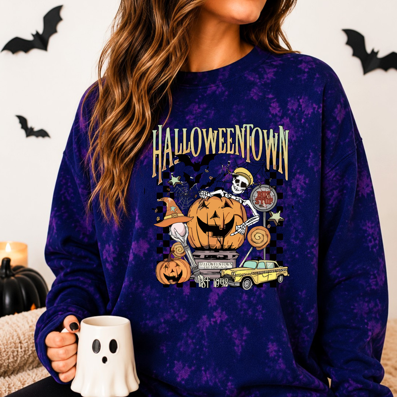 A woman wearing a purple and navy blue tie-dye sweatshirt with a colorful 'Halloween Town' graphic featuring pumpkins, a skeleton, a ghost, and a yellow car. She is holding a white ghost-shaped mug in a cozy indoor setting.