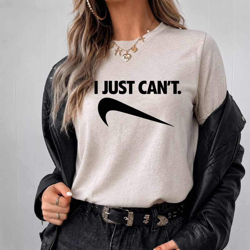 A woman wearing a light beige heather crewneck sweatshirt with the black text 'I JUST CAN'T.' and a black NKE swoosh graphic.