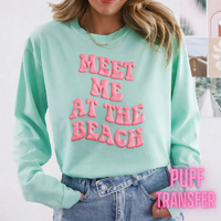 Person wearing a light blue sweatshirt with puff pink text 'MEET ME AT THE BEACH' and denim shorts.